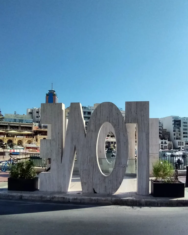 A street-level daytime view of the travertine stone Love Monument in St. Julian's, Malta, featuring large inverted and backward letters spelling the word LOVE, with buildings and a bright blue sky in the background.